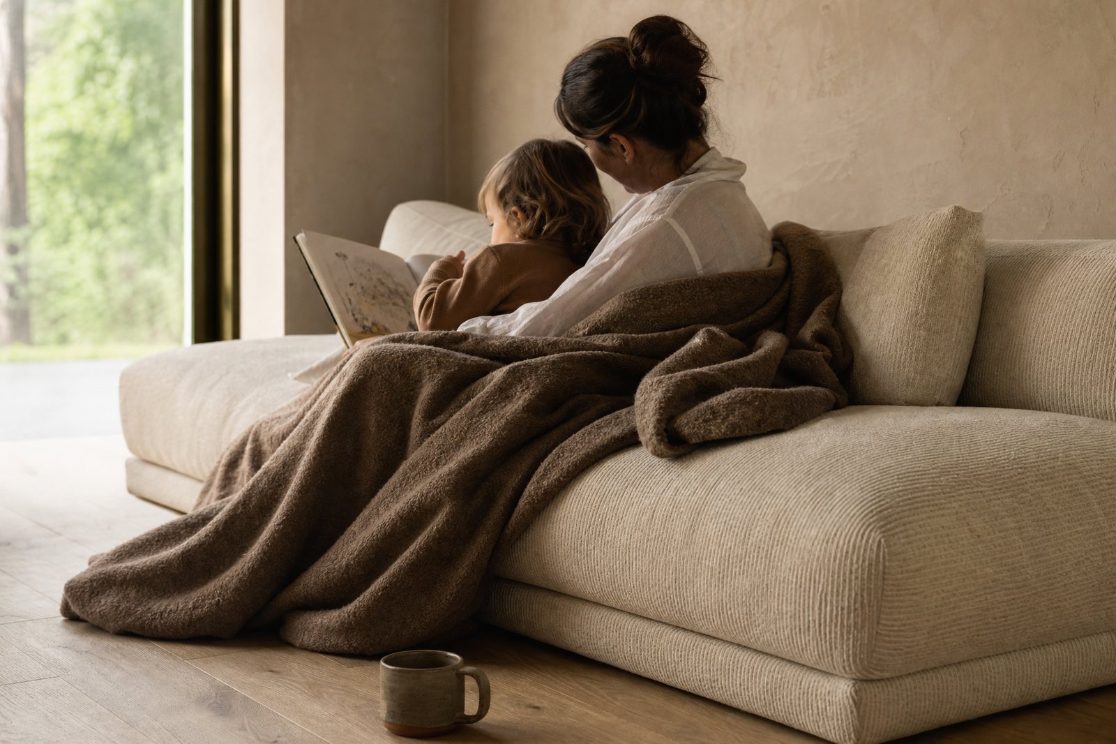 A parent and child reading under a brown wool throw on an oatmeal sofa